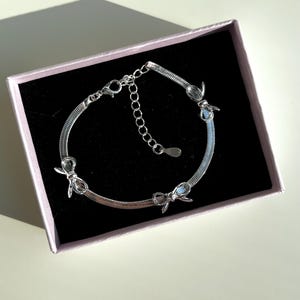 May include: A silver bracelet with three bow-shaped charms. The bracelet is in a white box with a black interior.