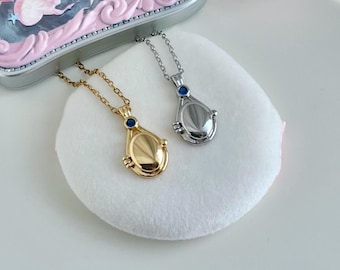 May include: Two teardrop-shaped pendant necklaces, one gold-toned and one silver-toned, each with a small blue gem. The pendants rest on a white, fluffy surface, with a decorative, ornate mirror in the background. The necklaces are on delicate chains.