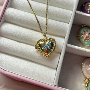 Puede incluir: Collar con colgante en forma de corazón dorado con una fina cadena. El medallón presenta un diseño de abeja azul y dorada. El collar se exhibe en una caja de joyería rosa con otros colgantes decorativos.