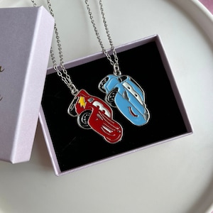 May include: Two silver chains with pendants of cartoon cars. One car is red with a yellow lightning bolt and the other is blue. The chains are in a light purple box with a black interior.