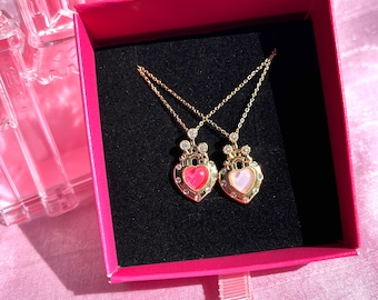 May include: Two gold necklaces with heart-shaped pendants. The pendants are adorned with rhinestones and have a crown design. One pendant is pink and the other is red. The necklaces are displayed in a pink box.