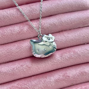 Puede incluir: Collar plateado con un colgante con forma de gato de dibujos animados con un lazo. El colgante tiene una superficie brillante y está sujeto a una cadena fina. El collar se presenta sobre una superficie rosa y aterciopelada.