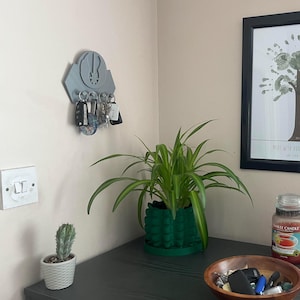 May include: A dark grey key holder with a decorative design and keys hanging from it. A green potted plant, a cactus in a white pot, two red glass candle jars, and a wooden bowl with keys and a remote control are on a dark grey surface.