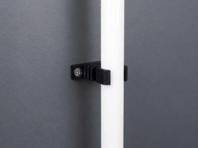 K&ouml;nnte beinhalten: Ein schwarzer Kunststoff-Wandclip mit geripptem Design und einer einzelnen silberfarbenen Schraube, der eine glatte, vertikale wei&szlig;e zylindrische Stange fest gegen eine dunkelgraue Oberfl&auml;che h&auml;lt.