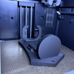 K&ouml;nnte beinhalten: Ein mattschwarzer 3D-gedruckter St&auml;nder mit einer kreisf&ouml;rmigen Basis, einer vertikalen scheibenf&ouml;rmigen Komponente und einer U-f&ouml;rmigen Halterung. Das Objekt befindet sich auf der texturierten Druckplatte eines 3D-Druckers, wobei der Druckkopf dar&uuml;ber sichtbar ist.