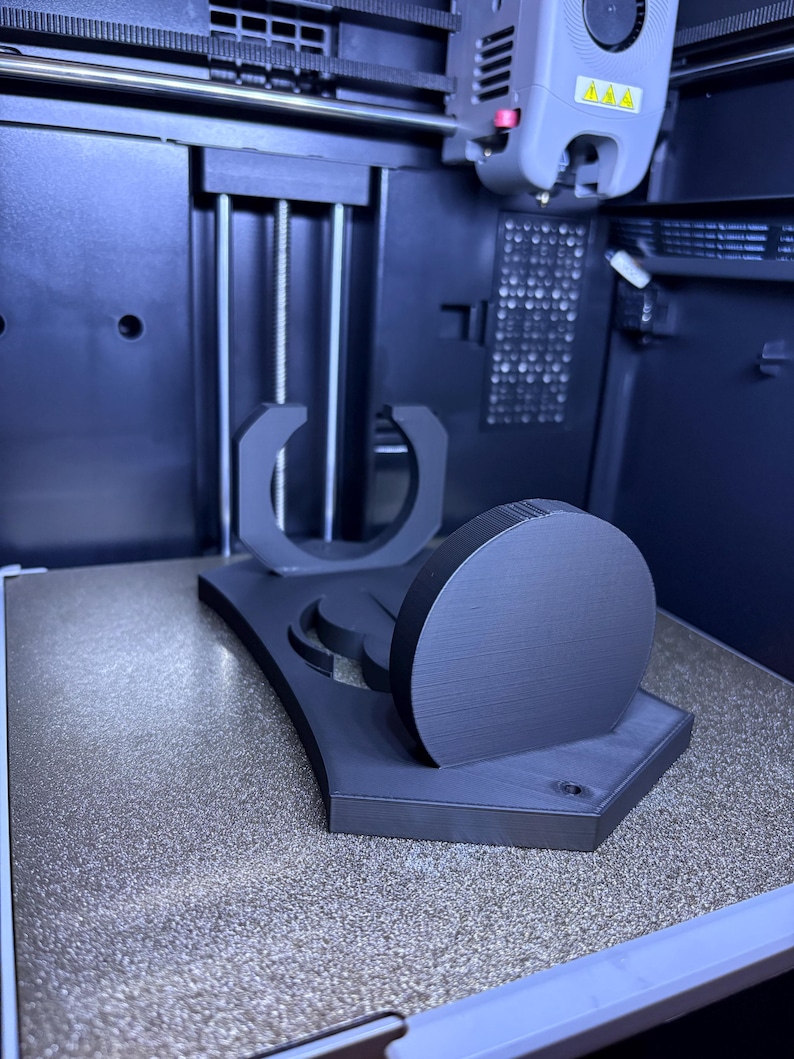 K&ouml;nnte beinhalten: Ein mattschwarzer 3D-gedruckter St&auml;nder mit einer kreisf&ouml;rmigen Basis, einer vertikalen scheibenf&ouml;rmigen Komponente und einer U-f&ouml;rmigen Halterung. Das Objekt befindet sich auf der texturierten Druckplatte eines 3D-Druckers, wobei der Druckkopf dar&uuml;ber sichtbar ist.