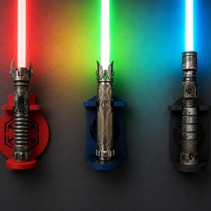 K&ouml;nnte beinhalten: Drei wandmontierte Lichtschwerter mit leuchtenden Klingen in Rot, Gr&uuml;n und Blau. Jeder Griff weist markante metallische Details auf und wird von einer kreisf&ouml;rmigen Halterung in passenden Farben, Rot, Blau und Schwarz, vor dunklem Hintergrund gehalten.