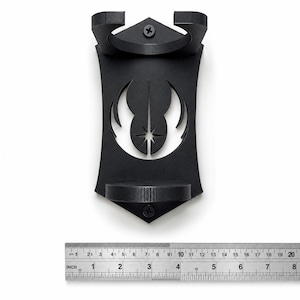 K&ouml;nnte beinhalten: Ein mattschwarzer Wandhalter mit einem Ausschnitt des Jedi-Orden-Symbols. Der Halter verf&uuml;gt &uuml;ber zwei kreisf&ouml;rmige Ringe zur St&uuml;tzung und ist mit zwei sichtbaren Schrauben befestigt. Ein darunter liegendes Metalllineal zeigt eine Breite von ca. 11,5 cm.