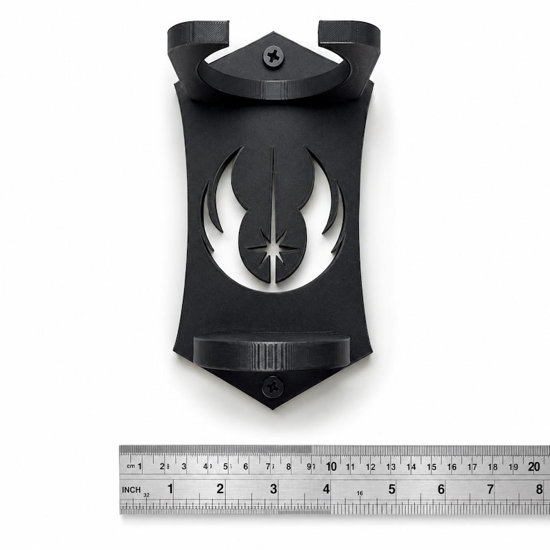 K&ouml;nnte beinhalten: Ein mattschwarzer Wandhalter mit einem Ausschnitt des Jedi-Orden-Symbols. Der Halter verf&uuml;gt &uuml;ber zwei kreisf&ouml;rmige Ringe zur St&uuml;tzung und ist mit zwei sichtbaren Schrauben befestigt. Ein darunter liegendes Metalllineal zeigt eine Breite von ca. 11,5 cm.