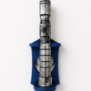 K&ouml;nnte beinhalten: Ein metallisch silberner und schwarzer, verwitterter Lichtschwertgriff, der vertikal an einer dunkelblauen Wandhalterung aus Kunststoff befestigt ist. Der Griff hat ein zylindrisches Design mit horizontalen Rillen und einem runden Schalter.