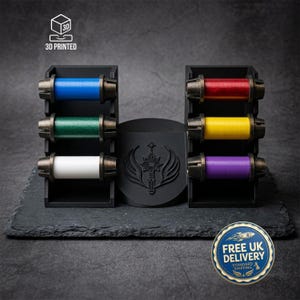 Könnte beinhalten: Ein schwarzer 3D-gedruckter Präsentationsständer mit zwei vertikalen Regalen, die sechs zylindrische Spulen in Blau, Grün, Weiß, Rot, Gelb und Lila halten. Ein zentrales schwarzes kreisförmiges Emblem zeigt ein Schwert- und Flügelmotiv. Der Text 3D PRINTED ist sichtbar.