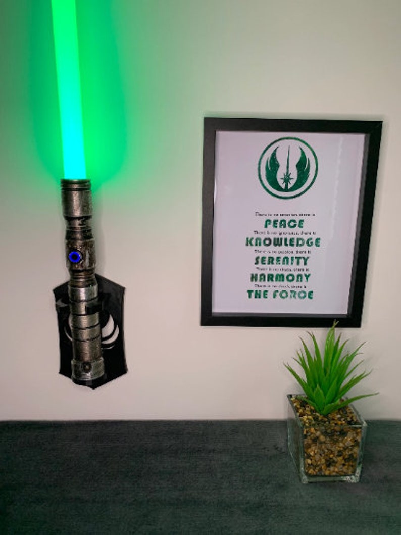 K&ouml;nnte beinhalten: Ein metallisch silbernes Lichtschwert mit leuchtend gr&uuml;ner Klinge, montiert an einer schwarzen Wandhalterung, neben einem gerahmten Druck mit gr&uuml;nem Jedi-Symbol und dem Text: There is no emotion, there is PEACE. There is no ignorance, there is KNOWLEDGE.