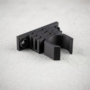K&ouml;nnte beinhalten: Eine schwarze 3D-gedruckte Montagehalterung mit einer flachen rechteckigen Basis, zwei kreisf&ouml;rmigen Schraubl&ouml;chern an den Enden und einer zentralen zylindrischen Klemme, die dazu dient, eine Stange oder ein Werkzeug sicher mit matter Oberfl&auml;che zu halten.