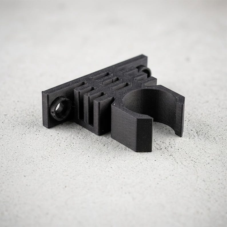 K&ouml;nnte beinhalten: Eine schwarze 3D-gedruckte Montagehalterung mit einer flachen rechteckigen Basis, zwei kreisf&ouml;rmigen Schraubl&ouml;chern an den Enden und einer zentralen zylindrischen Klemme, die dazu dient, eine Stange oder ein Werkzeug sicher mit matter Oberfl&auml;che zu halten.