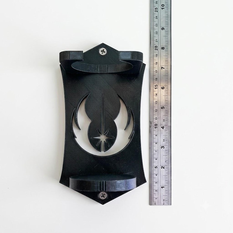 K&ouml;nnte beinhalten: Eine schwarze, 3D-gedruckte Wandhalterung mit einem mittigen Ausschnitt des Jedi-Ordens-Symbols. Die Halterung verf&uuml;gt &uuml;ber zwei kreisf&ouml;rmige Aufnahmen und ist mit zwei silbernen Schrauben befestigt. Ein Lineal zeigt eine L&auml;nge von ca. 26 cm.