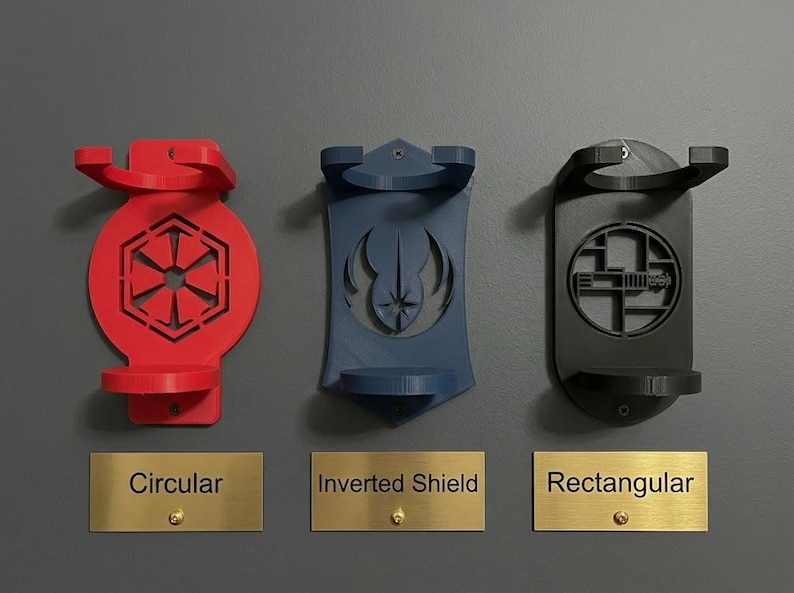 K&ouml;nnte beinhalten: Drei Wandhalterungen in Rot, Blau und Schwarz. Die rote Halterung ist kreisf&ouml;rmig mit einem sechseckigen Emblem, die blaue ist ein umgekehrtes Schild mit einem Symbol und die schwarze ist rechteckig mit einem geometrischen Muster. Die Schilder tragen die Aufschriften Circular, Inverted Shield, Rectangular.
