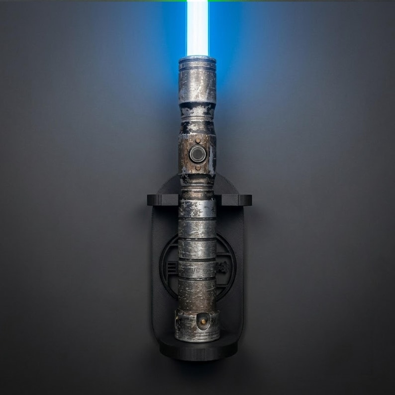K&ouml;nnte beinhalten: Ein verwitterter metallischer Lichtschwertgriff mit leuchtend blauer Klinge, vertikal montiert in einer schwarzen Wandhalterung. Der Griff hat eine texturierte, abgenutzte silberne Oberfl&auml;che mit horizontalen Rillen und einem runden Knopf.