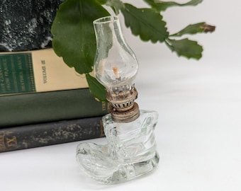 May include: A clear glass oil lamp shaped like a boot. The lamp has a small glass chimney and a metal base. The boot-shaped base is detailed with embossed lines. The lamp is sitting on a white surface.