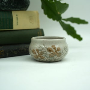 May include: A small, white ceramic bowl with a brown floral design. The bowl is sitting on a white surface.