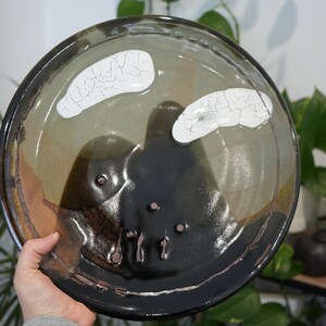 May include: A handcrafted ceramic plate with a distinctive design. The plate has a dark brown rim, a central area of black and olive green, and two white, cracked-patterned accents. The plate is round and appears hand-painted.
