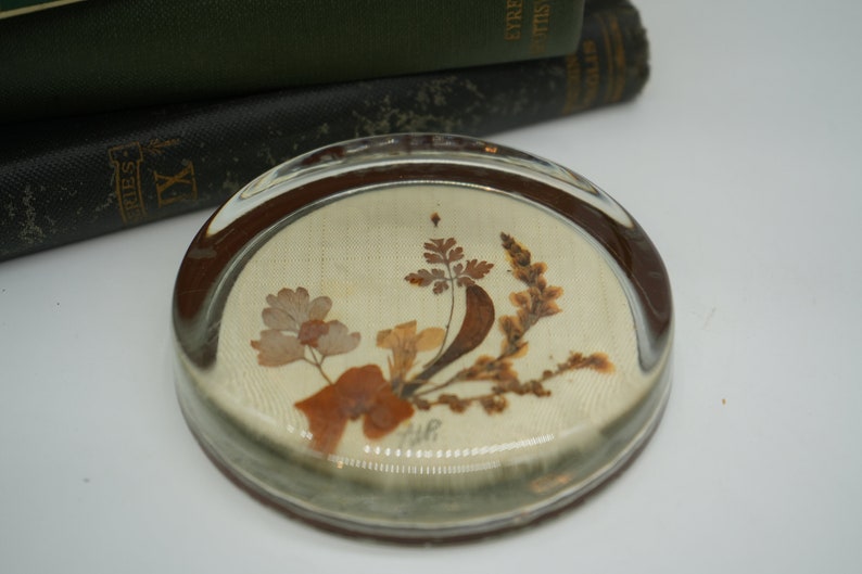 May include: A round glass paperweight with a pressed floral design. The design features dried flowers and leaves in shades of brown and beige on a white background.