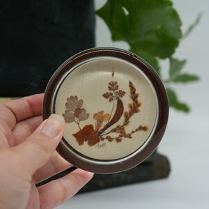 May include: A round, brown and cream-colored coaster with a pressed flower design. The coaster features a floral arrangement of dried leaves and flowers in shades of brown, cream, and beige.
