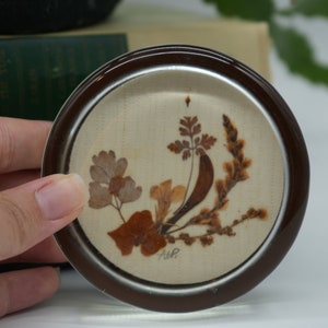 May include: A round glass coaster with a brown rim and a white background. The coaster features a pressed flower design with brown and tan leaves and flowers. The coaster is signed "A.W.P."