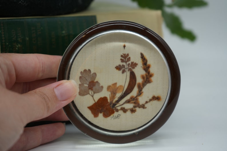 May include: A round glass coaster with a brown rim and a white background. The coaster features a pressed flower design with brown and tan leaves and flowers. The coaster is signed "A.W.P."