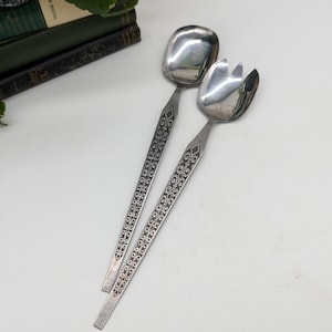 May include: A pair of vintage silver salad servers. One server has a square spoon-like head, the other has a fork-like head. Both have long handles with an etched design. The servers are displayed on a white surface with a stack of green books in the background.