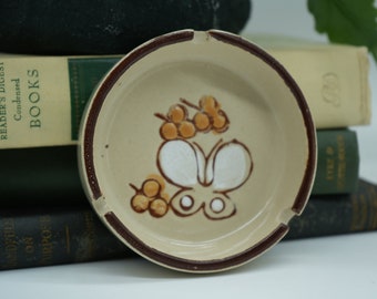 May include: A ceramic ashtray with a brown rim and a beige interior. The ashtray features a hand-painted design of a white butterfly with brown accents and yellow flowers.