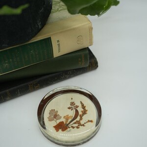 May include: A round glass paperweight with a pressed flower design. The paperweight has a clear glass base and a white background with brown and yellow pressed flowers.