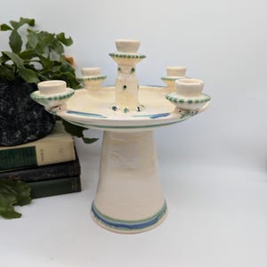 May include: A white ceramic candle holder with five candle cups. The holder has a round base and a tall, cylindrical stem. The candle cups are decorated with green and blue accents.