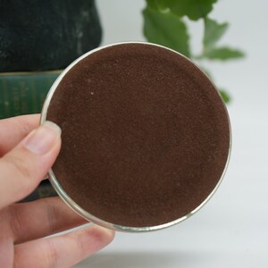 May include: A round, silver-colored coaster with a brown felt bottom. The coaster is being held in a hand.