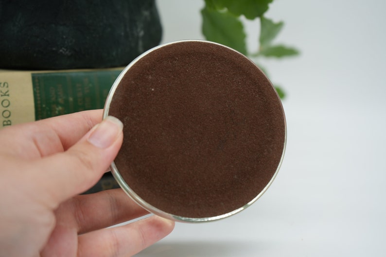 May include: A round, silver-colored coaster with a brown felt bottom. The coaster is being held in a hand.