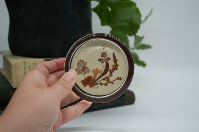 May include: A small, round, brown and white ceramic coaster with a pressed flower design. The coaster features a brown rim and a white center with a pressed flower design of brown and tan leaves and stems.