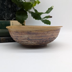 May include: A handcrafted ceramic bowl with a textured surface. The bowl has a wide, shallow shape and is finished in earthy tones of brown and tan. The exterior has a rough, organic texture, while the interior is smooth. A unique piece for serving or display, approximately 25cm in diameter.