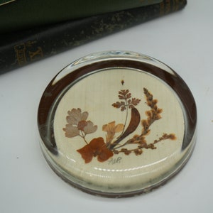 May include: A round glass paperweight with a brown rim and a white background. The paperweight features a pressed flower design with brown and beige flowers and leaves. The text "AWR" is visible in the center of the paperweight.