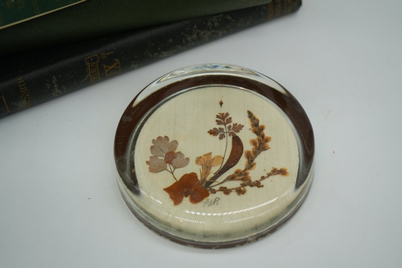 May include: A round glass paperweight with a brown rim and a white background. The paperweight features a pressed flower design with brown and beige flowers and leaves. The text "AWR" is visible in the center of the paperweight.