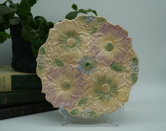 Op de afbeelding: Een decoratief keramiek bord met een reliëf bloemmotief. Het bord heeft een lichtgele achtergrond met roze, groene en blauwe bloemen.