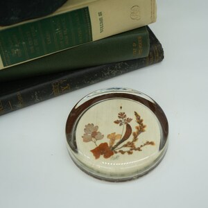 May include: A round glass paperweight with a beige background and a pressed flower design in shades of brown and beige.