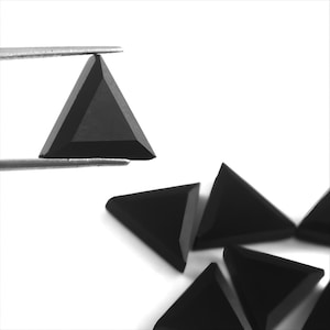 Könnte beinhalten: Nahaufnahme von mehreren schwarzen, dreieckigen Edelsteinen. Einer wird von einer Pinzette gehalten, die seine Facetten zeigt. Andere sind auf einer weißen Oberfläche verstreut, was ihre geometrische Form und dunkle Farbe hervorhebt.