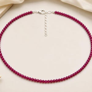 May include: A necklace composed of small, round, deep magenta beads arranged in a single strand. The necklace features a silver-toned metal lobster claw clasp and an adjustable chain extender with circular links at the back for a secure fit.