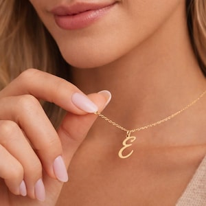 Könnte beinhalten: Eine goldfarbene Halskette mit einer filigranen Kette und einem Anhänger mit dem kursiven Buchstaben "E". Der Anhänger ist ein schlichtes, elegantes Design. Die Halskette wird von einer Hand mit gepflegten Nägeln gehalten.