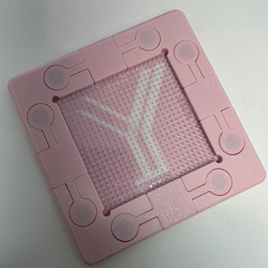 May include: A pink plastic puzzle with a white Y-shaped design in the center. The puzzle is made up of multiple pieces that fit together to form a square.