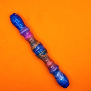 Può includere: Una bacchetta colorata e artigianale con un design a spirale. La bacchetta presenta sfumature di blu, rosa e oro, con una finitura glitterata. La bacchetta è posta su uno sfondo arancione brillante.