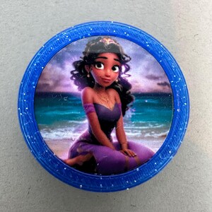 Puede incluir: Un botón redondo, azul, cubierto de purpurina con una imagen de una princesa de dibujos animados con cabello castaño y un vestido morado. La princesa está sentada en una playa con un océano azul de fondo.