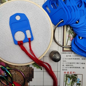 Könnte beinhalten: Nahaufnahme eines Kreuzstichprojekts mit einem blauen Plastik-Nadelfäder, rotem Stickgarn, einem hölzernen Stickrahmen, einem Fingerhut und einer Stickerschere. Das Projekt ist mit "H105 绿竹 Green bamb" beschriftet.