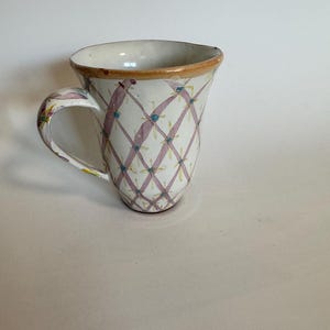 May include: A white ceramic mug with a handle, featuring a pink and green diamond pattern. The rim is trimmed with a brown band. The mug has a unique, slightly flared shape.