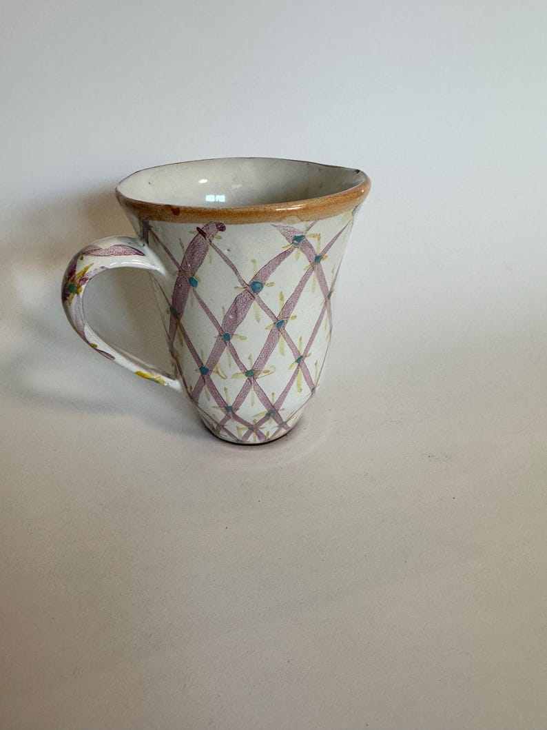 May include: A white ceramic mug with a handle, featuring a pink and green diamond pattern. The rim is trimmed with a brown band. The mug has a unique, slightly flared shape.
