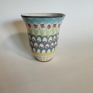 Op de afbeelding: Een keramische vaas met een uitlopende bovenkant en een patroon. De vaas heeft een lichtblauwe rand, een herhalend patroon van rode tulpen en groene bladeren en een geel en wit geblokte basis. De vaas is ongeveer 15 cm hoog.